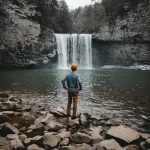 man standing on brown stone facing waterfalls